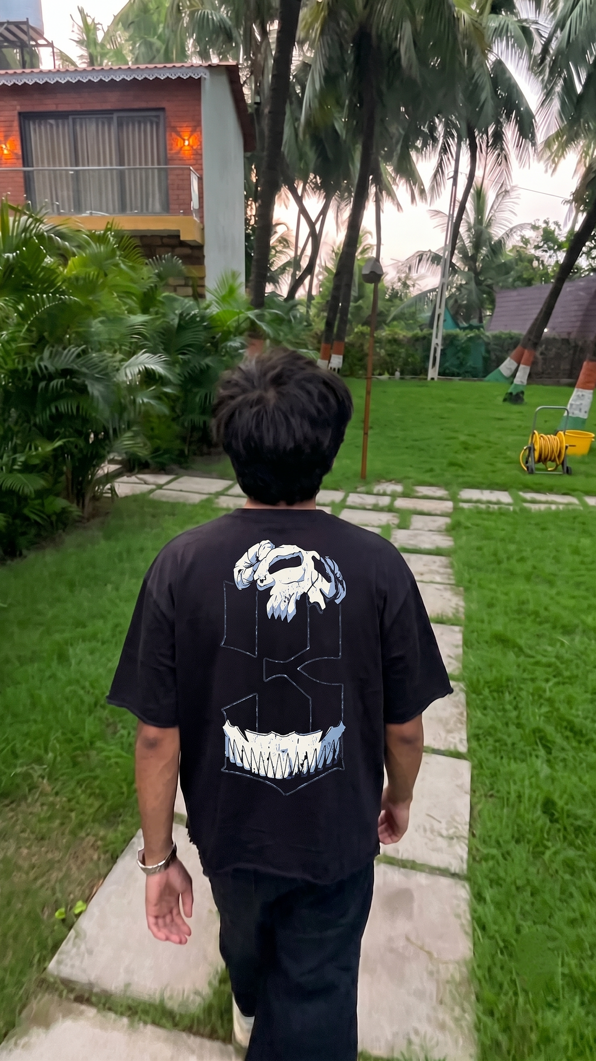 A man viewed from behind walking on a stone path in a grassy backyard, wearing a Scrollfit black t-shirt with a large white graphic of a skull and teeth on the back.