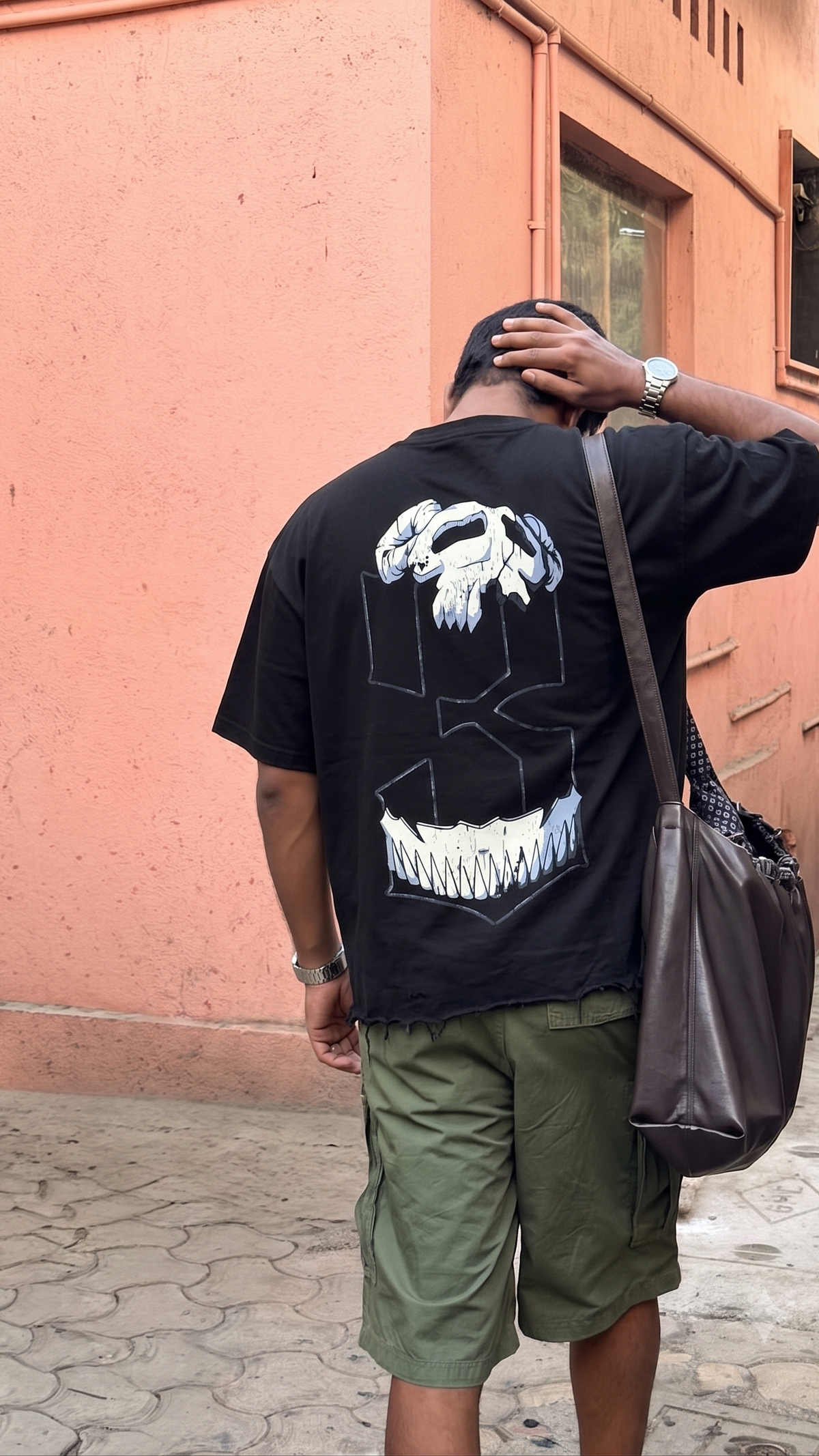A man viewed from behind wearing a black t-shirt with a Scrollfit large white graphic print on the back, paired with olive green cargo shorts and carrying a dark brown shoulder bag, standing outdoors against a peach-colored wall.
