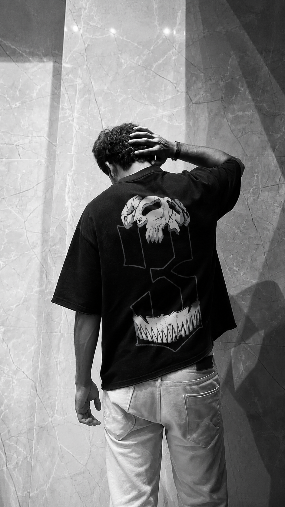 A man seen from behind wearing a Scrollfit black graphic t-shirt with a large white skull design on the back, paired with light-colored jeans, standing against a textured stone wall.