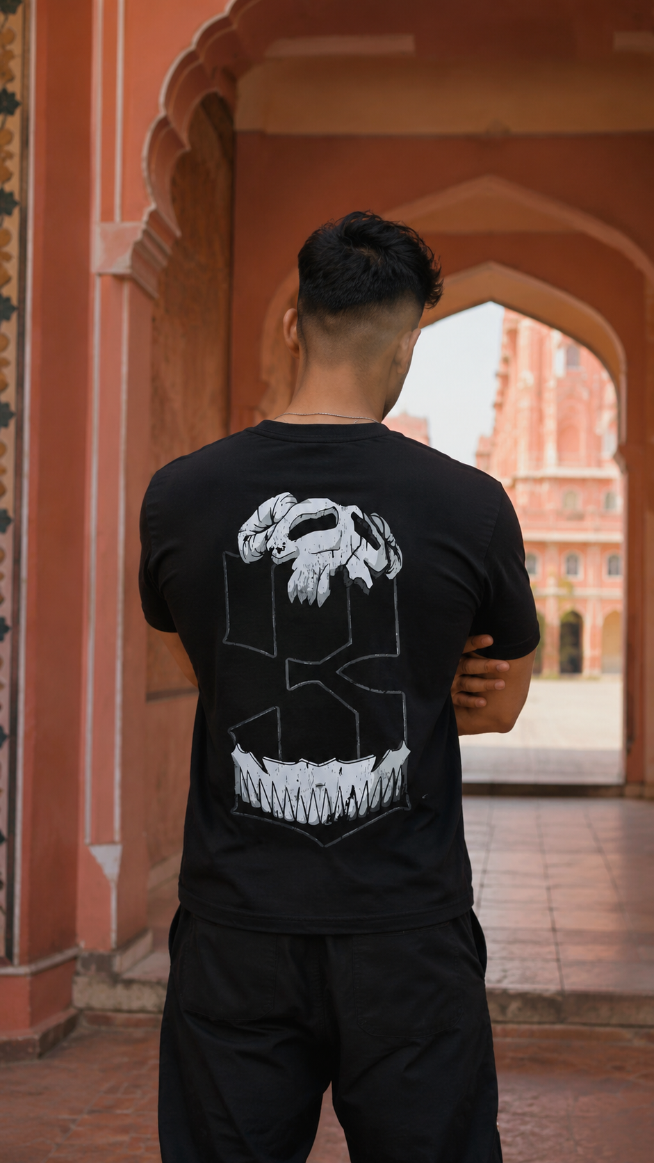 A man viewed from behind wearing a Scrollfit black t-shirt with a large white graphic of a skull inside a geometric frame, standing in front of a terracotta-colored arched corridor.