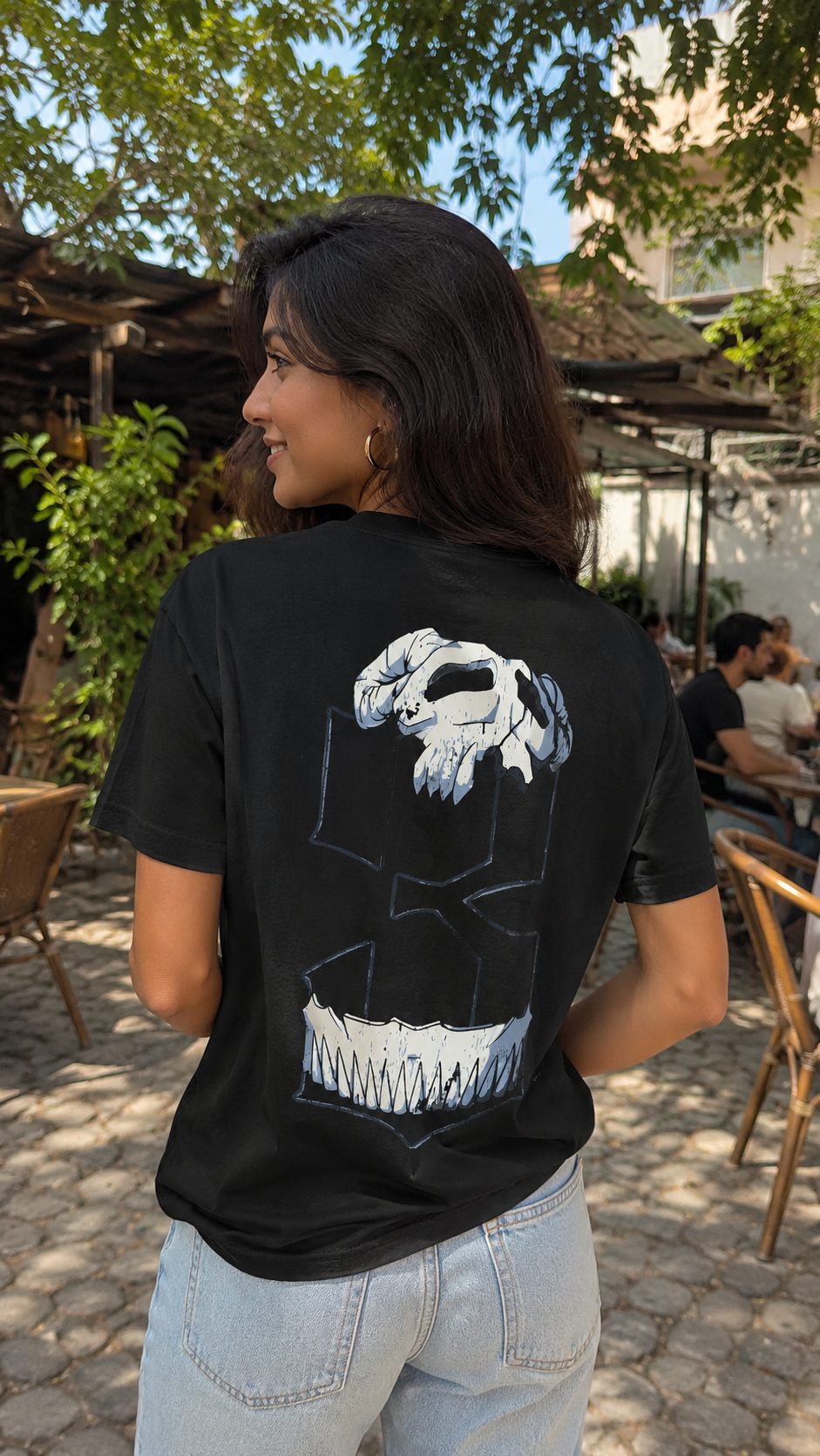 A woman wearing a Scrollfit black t-shirt with a large white and gray graphic print on the back, standing in an outdoor patio setting.