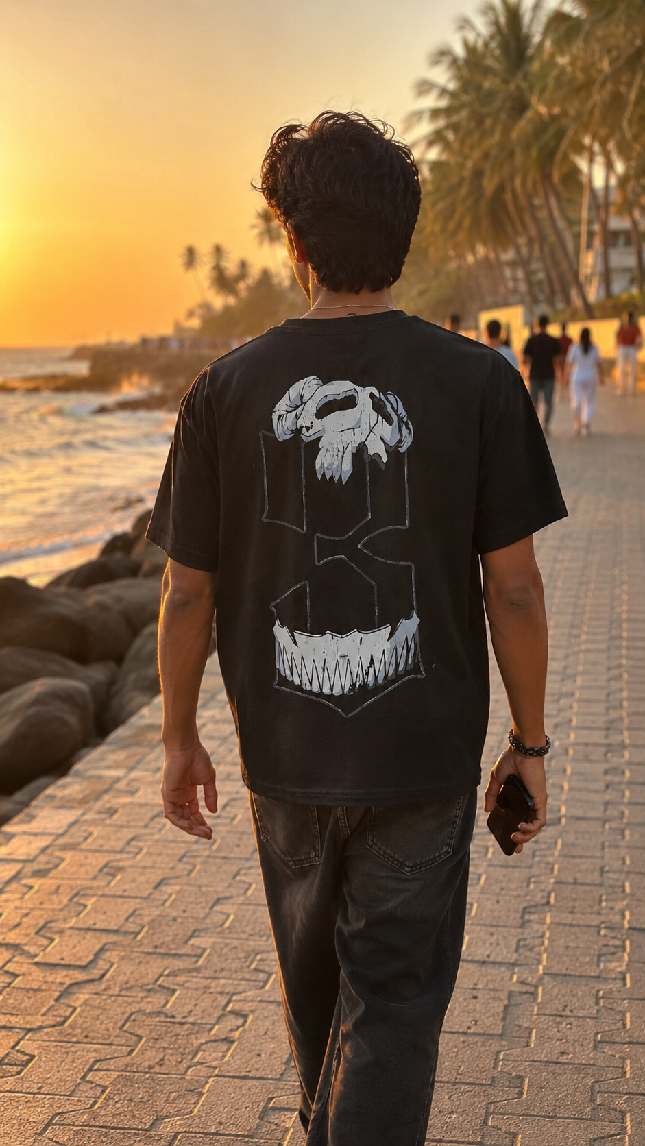 A man viewed from behind walking on a beach boardwalk at sunset, wearing a Scrollfit black graphic t-shirt with a large white shark design on the back and dark jeans.