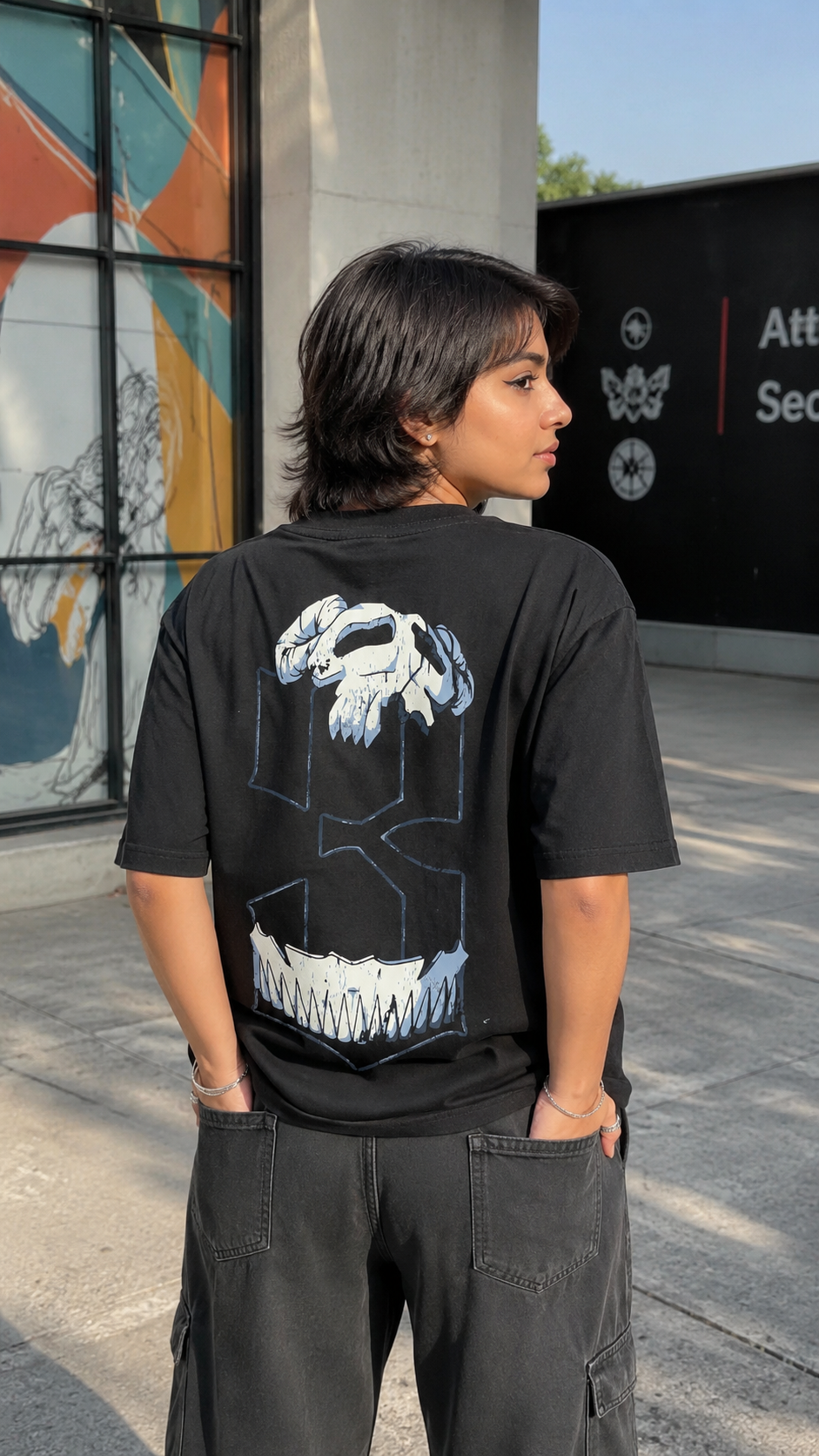 A person standing outdoors wearing a Scrollfit black graphic T-shirt with a large white and gray skull-and-shield design on the back, paired with dark cargo pants.