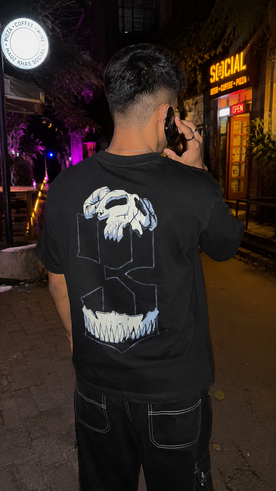 A man viewed from behind wearing a Scrollfit black t-shirt with a large white graphic of a skull inside a geometric frame on the back, standing on a city street at night.