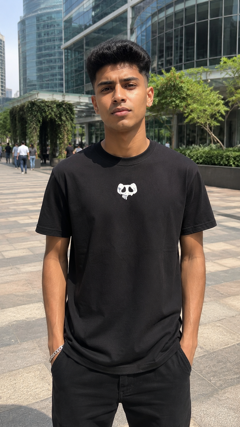 A young man wearing a Scrollfit black crewneck t-shirt with a small white skull graphic on the chest, standing outdoors in front of modern glass buildings.