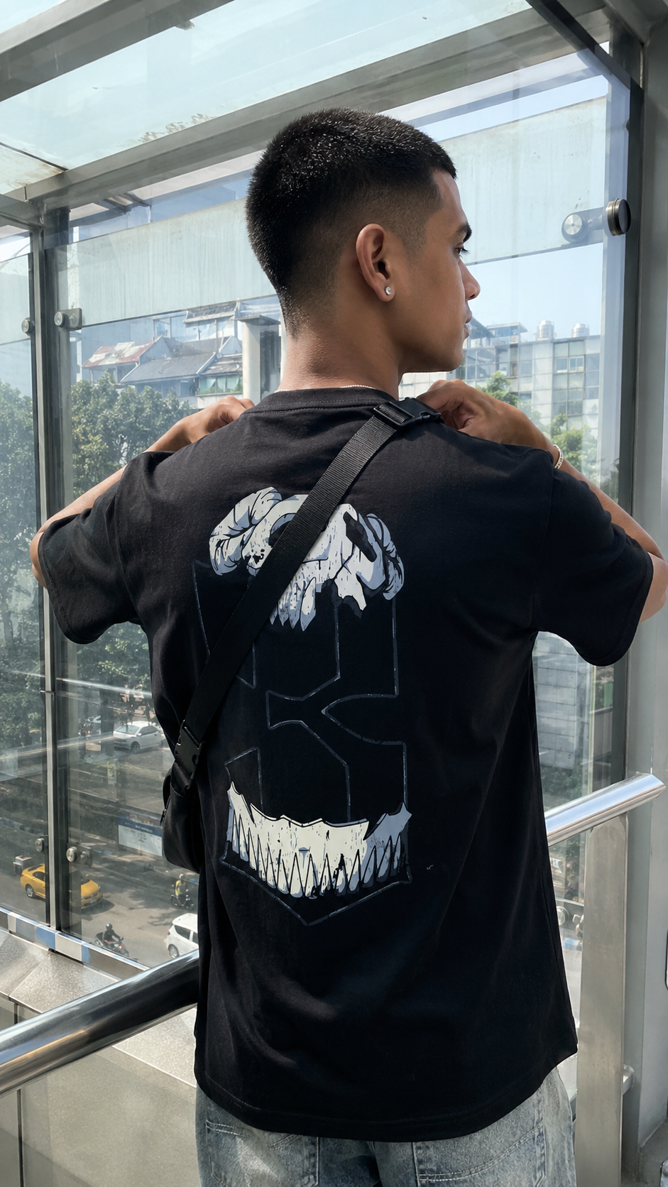 A man wearing a Scrollfit black t-shirt with a large white graphic print on the back, standing by a glass railing and looking out onto a city view.