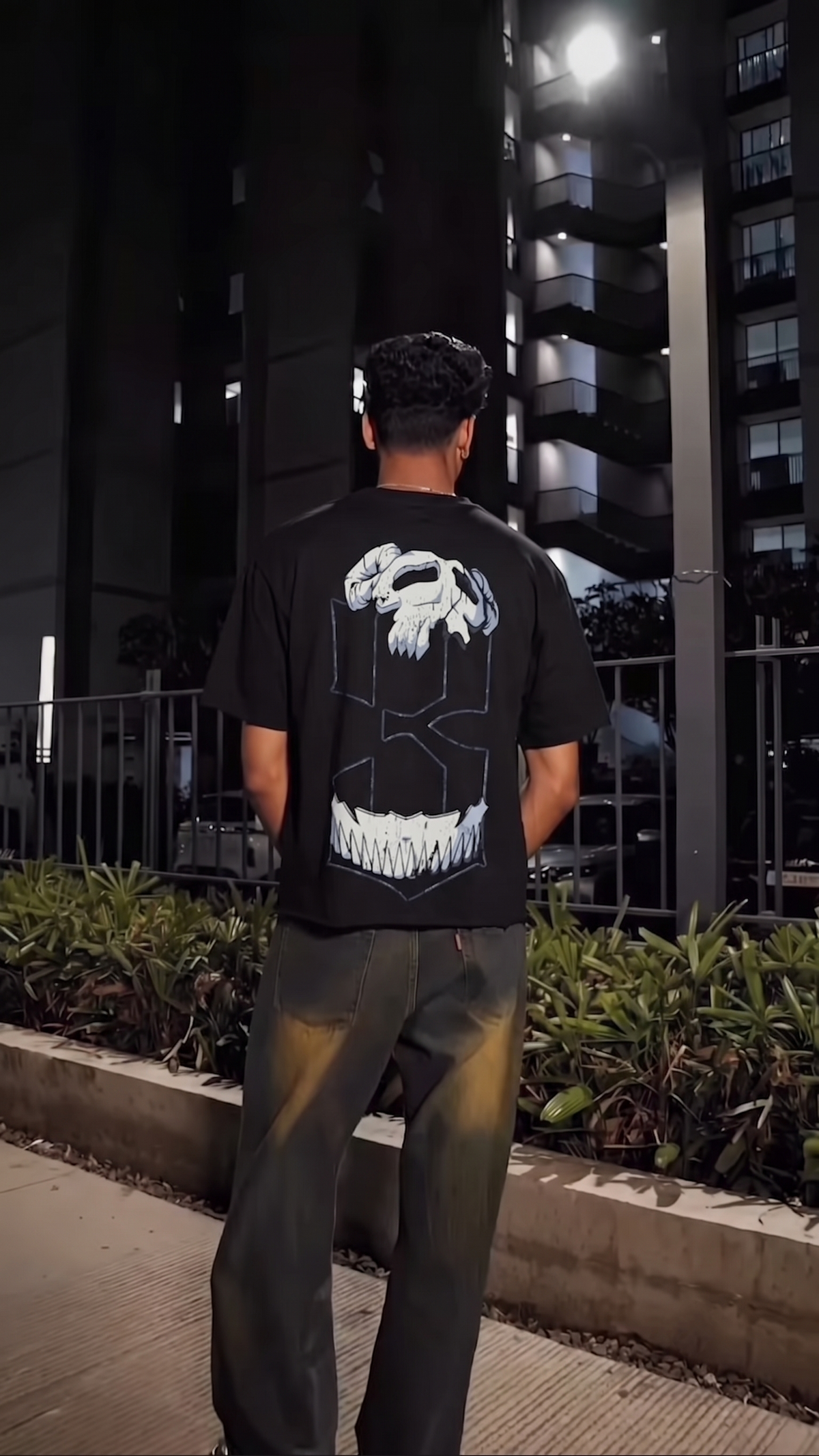 A man standing outdoors at night wearing a Scrollfit black graphic t-shirt with a large white skull design on the back, paired with dark pants.