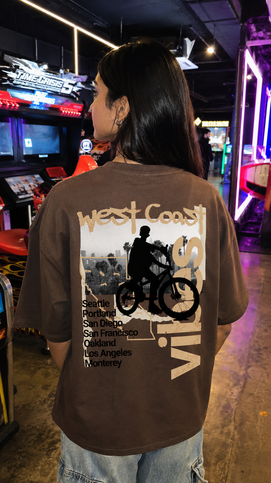 A person viewed from behind wearing a Scrollfit brown graphic t-shirt with a 'West Coast Vibes' bicycle design in an arcade setting.