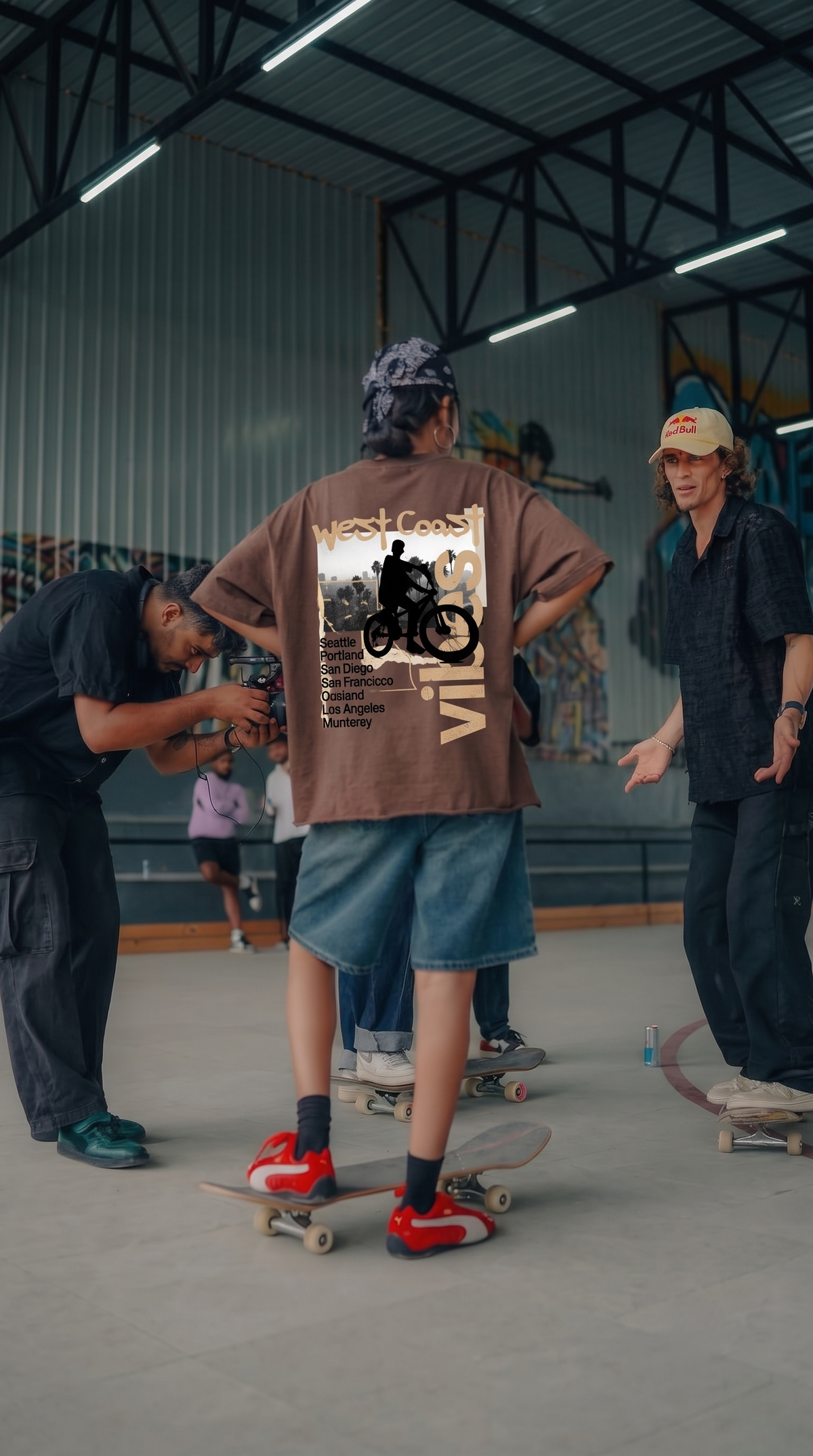 A person standing on a skateboard in an indoor skatepark, wearing a Scrollfit brown graphic T-shirt with a large back print, denim shorts, and red sneakers. Other people and skateboards are visible in the background.
