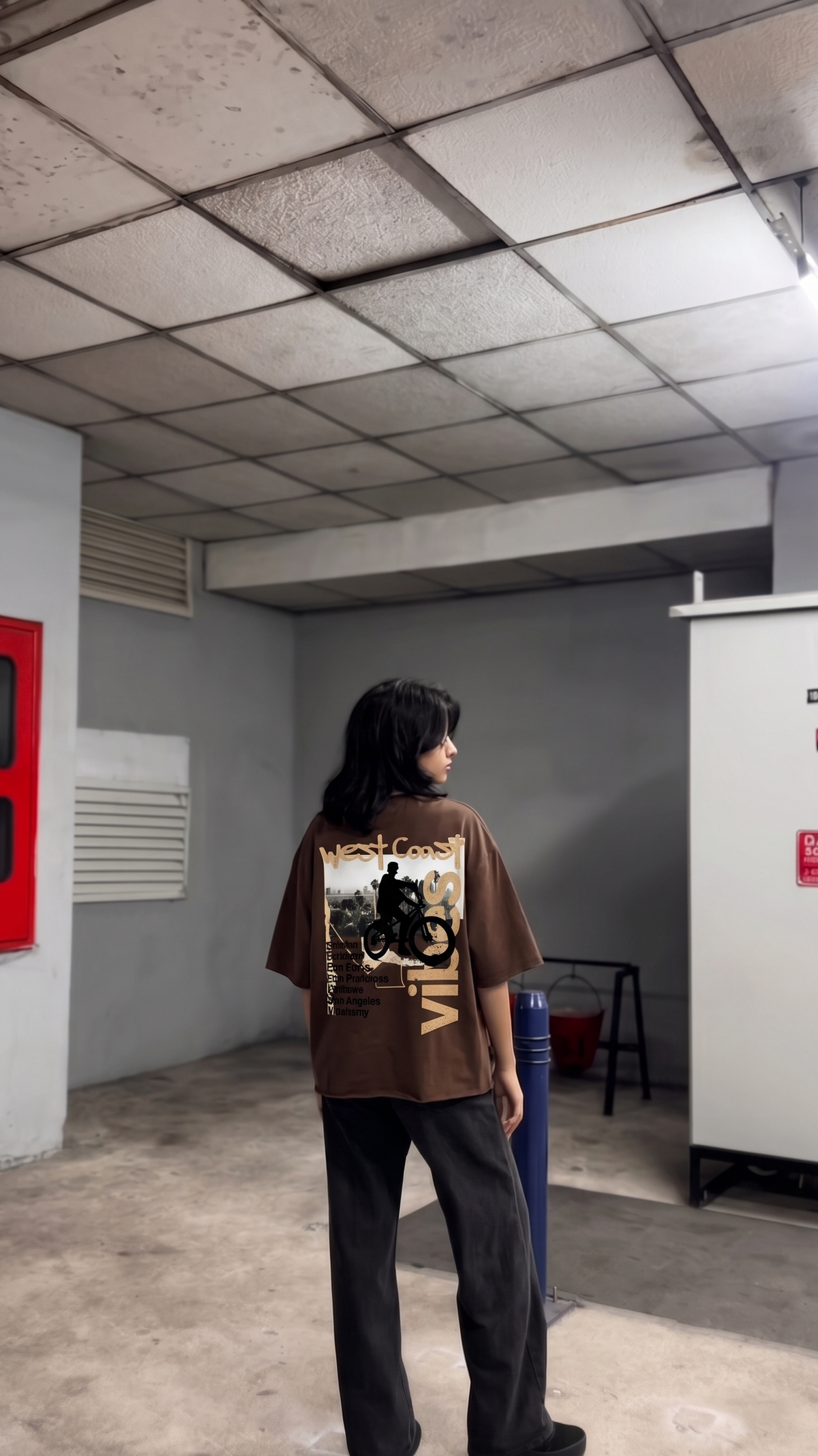 A person standing in a concrete indoor space wearing an oversized Scrollfit brown graphic T-shirt with a large motorcycle print on the back, paired with dark wide-leg pants.