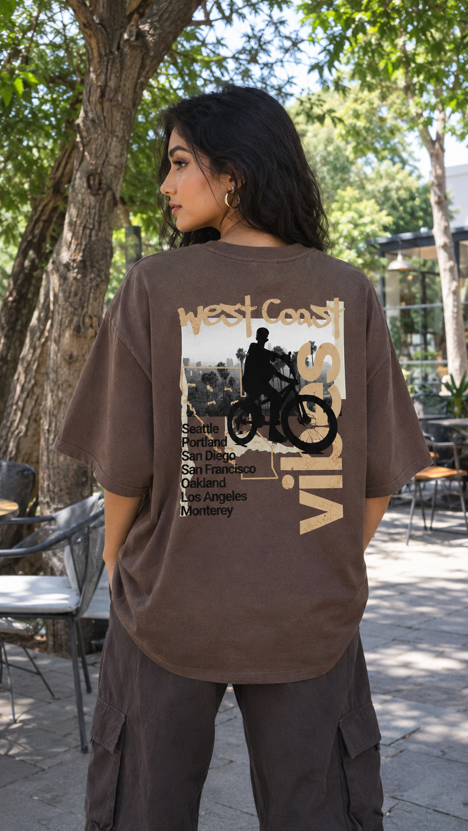 A woman wearing a Scrollfit brown oversized graphic t-shirt with a vintage-style print of a motorcyclist and the text 'West Coast Vibes', photographed outdoors in a park-like setting with trees and patio furniture.