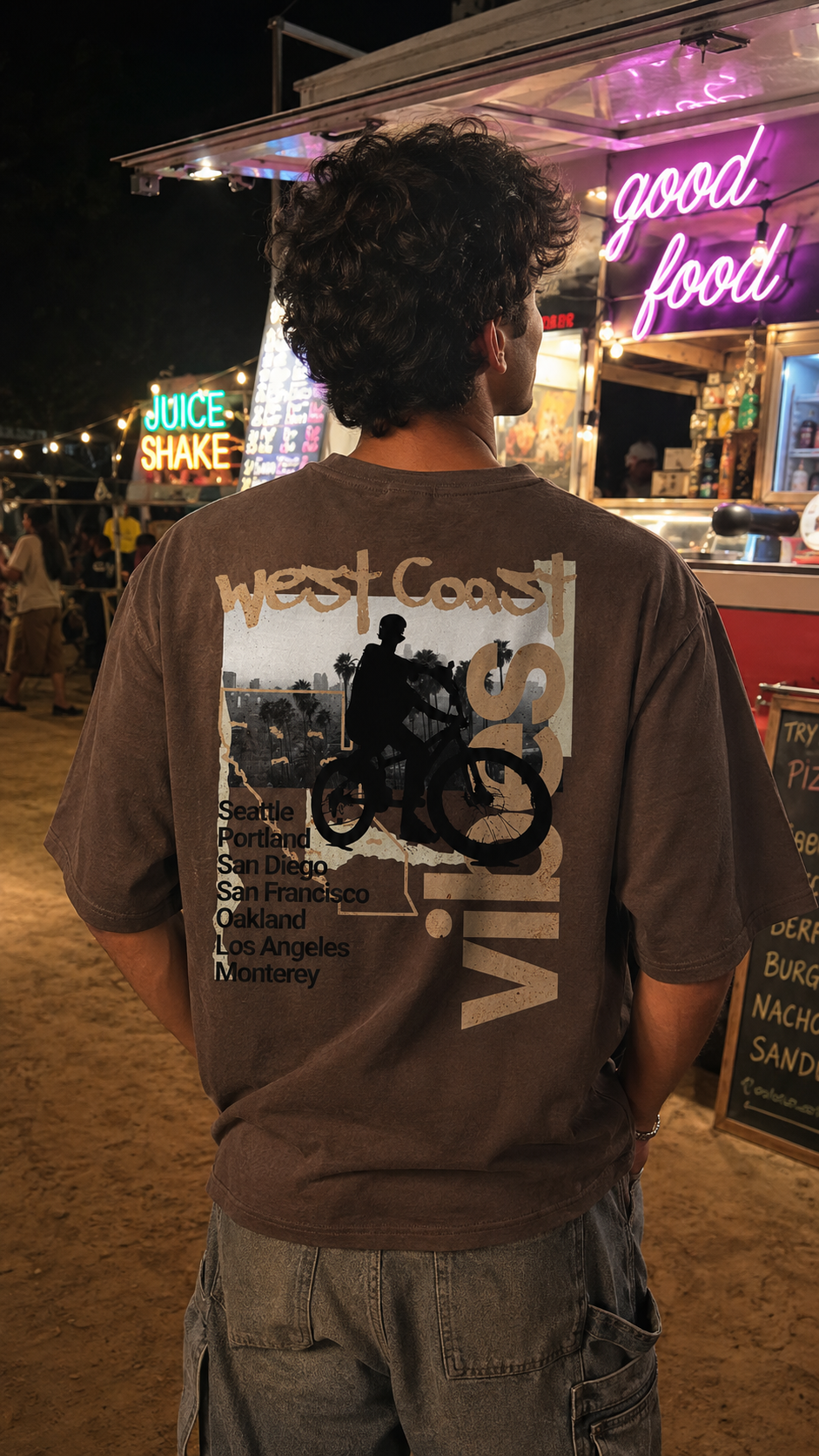 A person viewed from behind wearing a Scrollfit brown graphic t-shirt with a 'West Coast Vibes' motorcycle print, standing at night in front of a food stall with neon signs.