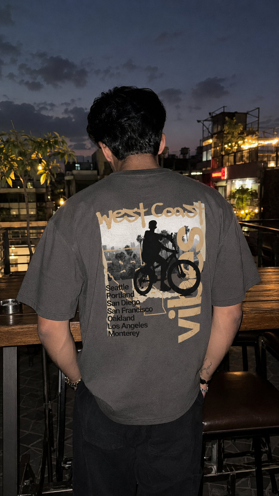 A man viewed from behind wearing a dark gray graphic t-shirt with a large Scrollfit 'West Coast Vibes' bicycle print on the back, set in an outdoor bar environment at dusk.