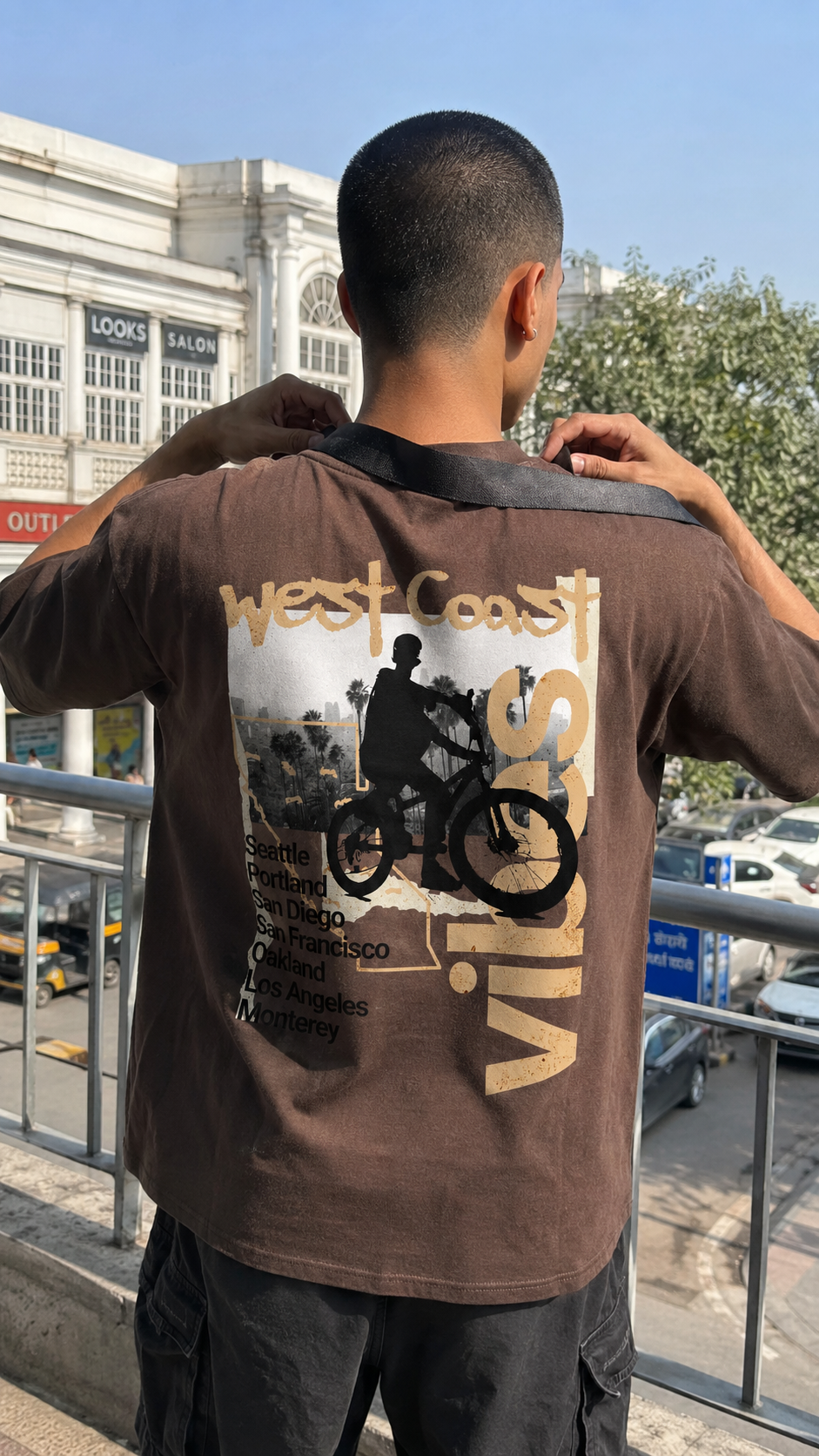 A man viewed from behind wearing a Scrollfit brown graphic t-shirt with a large back print featuring a motorcycle rider and the words 'west Coast' and 'vintage', set against an outdoor urban background.