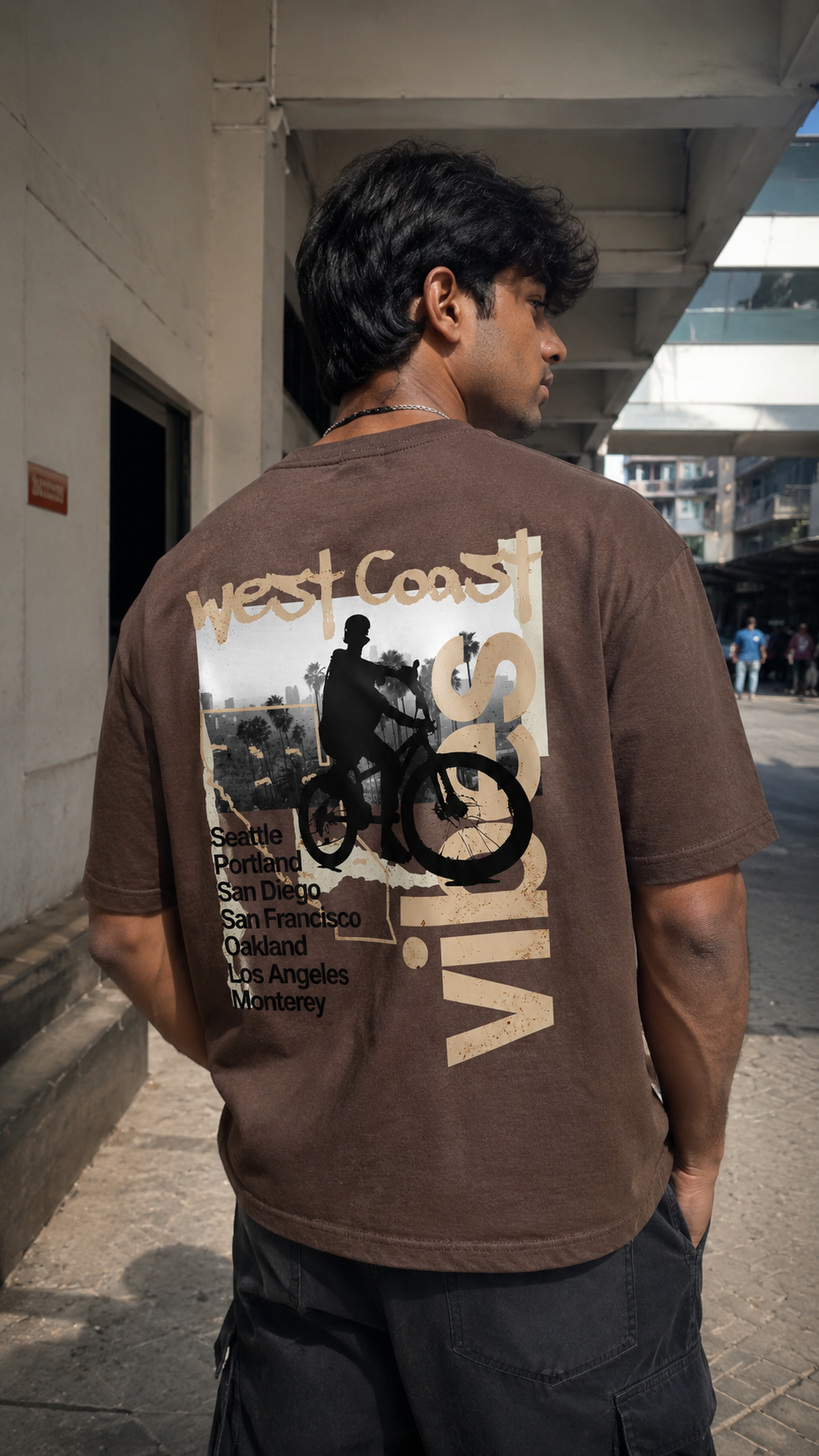 A man wearing a Scrollfit brown graphic t-shirt with a bicycle design and 'West Coast Vibes' text, photographed from behind in an outdoor urban setting.