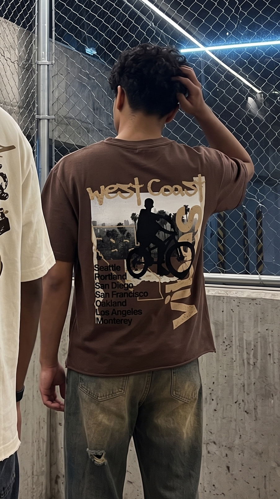 A person viewed from behind wearing a Scrollfit brown graphic T-shirt with a large back print reading 'West Coast' and listing coastal cities, paired with distressed jeans in an indoor industrial setting with a chain-link fence.