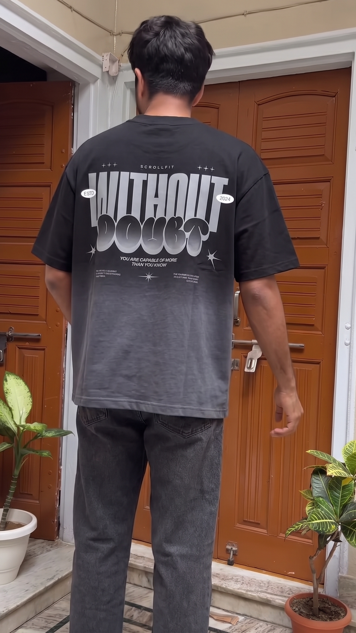 A man viewed from behind wearing a Scrollfit faded black graphic t-shirt with 'WITHOUT DOUBT' printed in white across the back, paired with dark gray jeans, standing in front of a brown wooden door.