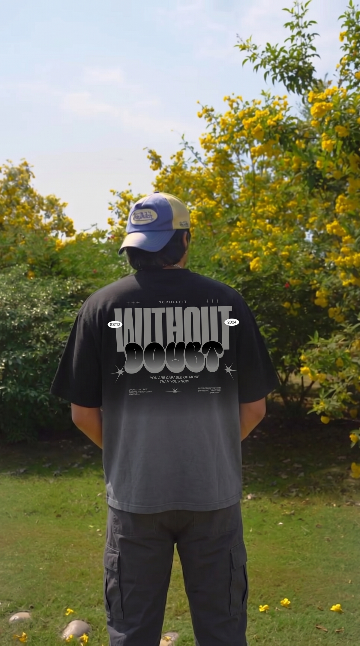 A man viewed from behind wearing a Scrollfit black t-shirt with a large white graphic print that reads 'WITHOUT DOUBT' on the back, paired with a blue and yellow trucker hat, standing in a grassy outdoor area with yellow flowering bushes.