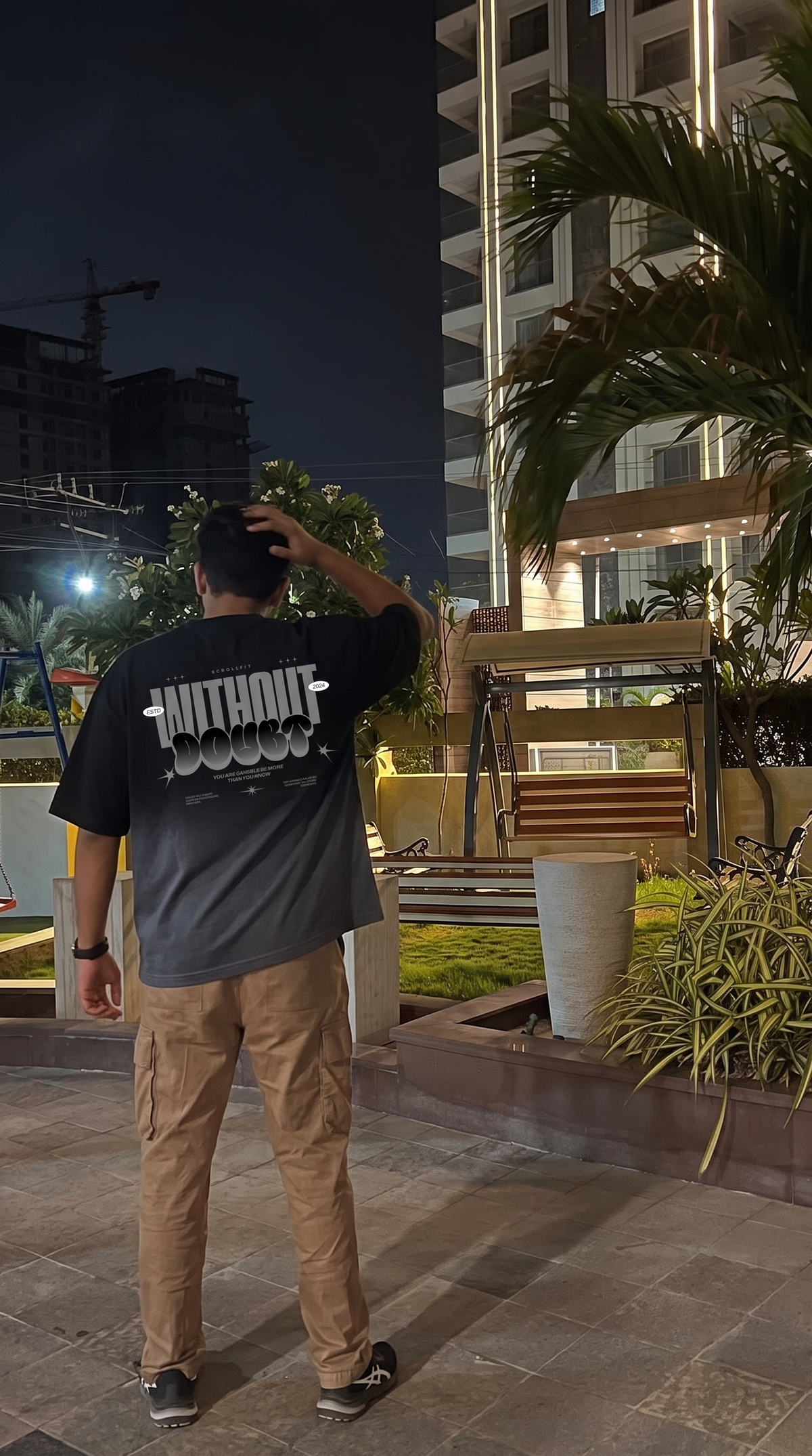 A man standing outdoors at night wearing a Scrollfit black graphic t-shirt with 'WITHOUT DOUBT' printed in white on the back, paired with tan cargo pants.