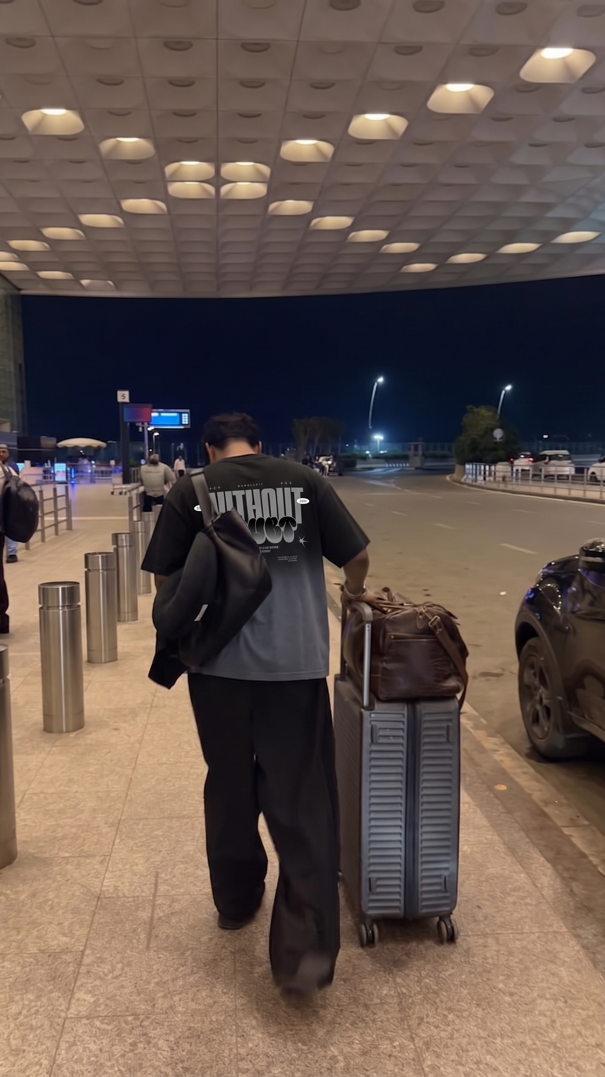A man walking away in an airport terminal at night, wearing a Scrollfit black t-shirt with a graphic print and carrying a black jacket over his shoulder while pulling a gray suitcase.